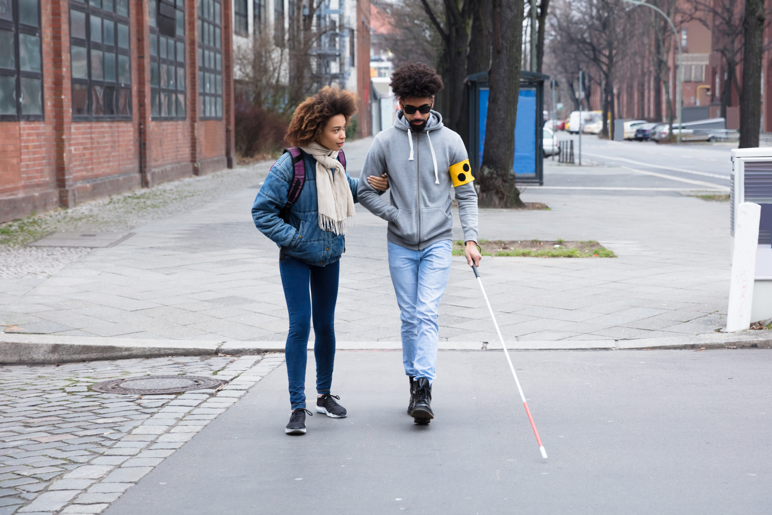a young man using a walking stick to cross the road with a young woman guiding him