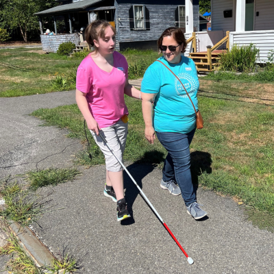 a staff member helps a teen with a white cane walk down the sidewalk