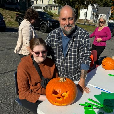 A girl with dark glasses and a man pose at a table with a jack-o-lantern on a beautiful fall day.