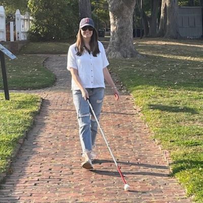 A young woman with a white cane walks down a brick sidewalk on a sunny day.