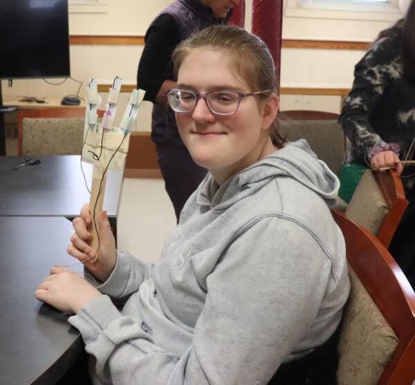 A young woman attending a workshop and wearing glasses holds up a hand-shaped craft she has created.