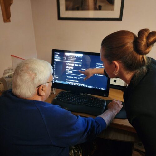 A man sits at a desktop computer as a woman points something out to him on the screen.