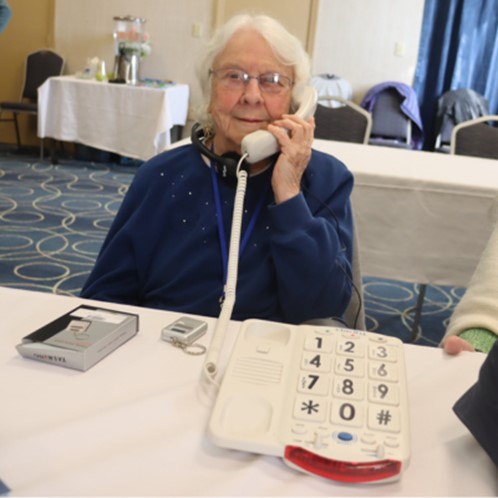 An older woman holds a Clarity J V 3 5 W phone to hear ear. The large buttons on the phone are visible on the table she sits at.