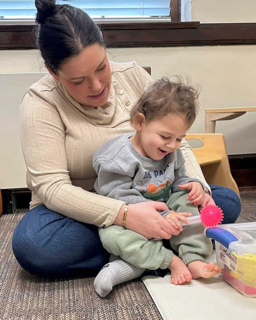 A teacher and toddler sit on a play mat with toys.