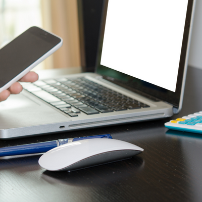 A desk with a laptop and mouse on it. A hand holds a smart phone.