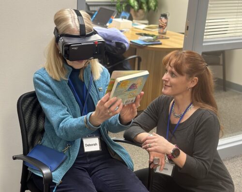 A senior woman wears V R glasses to look at a book while an instructor explains the technology.