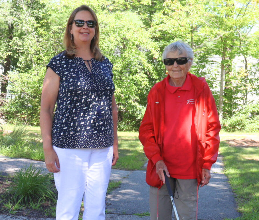 A senior woman stands with an Orientation & Mobility Specialist on a sidewalk in the shade of a tree.