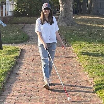 A young woman with a white cane walks on a brick walkway on a sunny day.