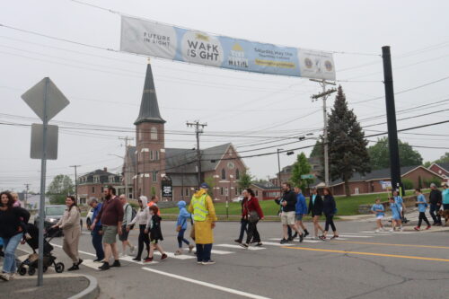 A group of people walk beneath a Walk For Sight banner spanning across a street.