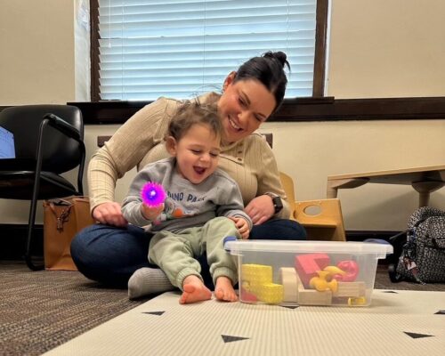 A woman sits on the floor and holds an infant in her lap who is holding a brightly colored toy.