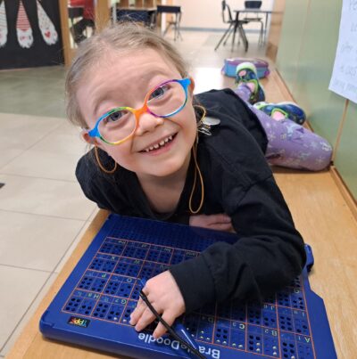 A young girl in glasses lies smiles as she does a braille activity.