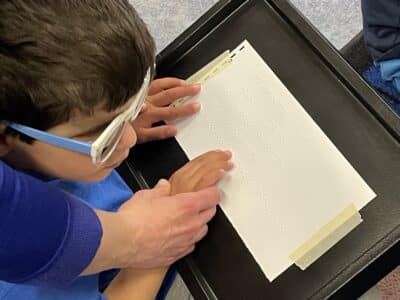A young boy being guided by a teacher reads braille.