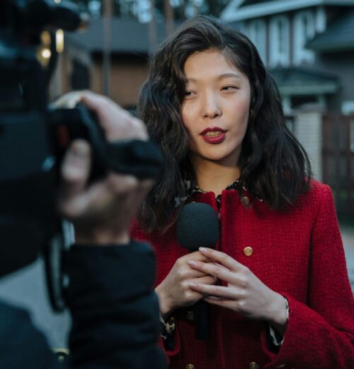 A female newscaster speaks into a microphone as a cameraman records her.