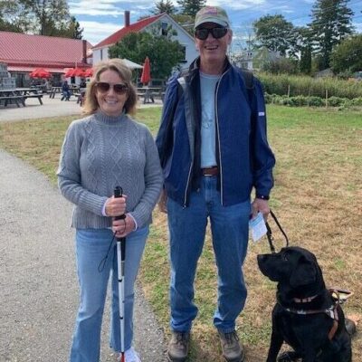 A woman with a white cane and a man with a guide dog pose together on a sunny day.