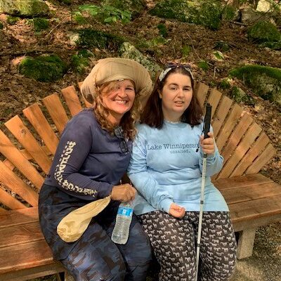 A woman and a teen girl with a white cane sit on a bench along a hiking trail.