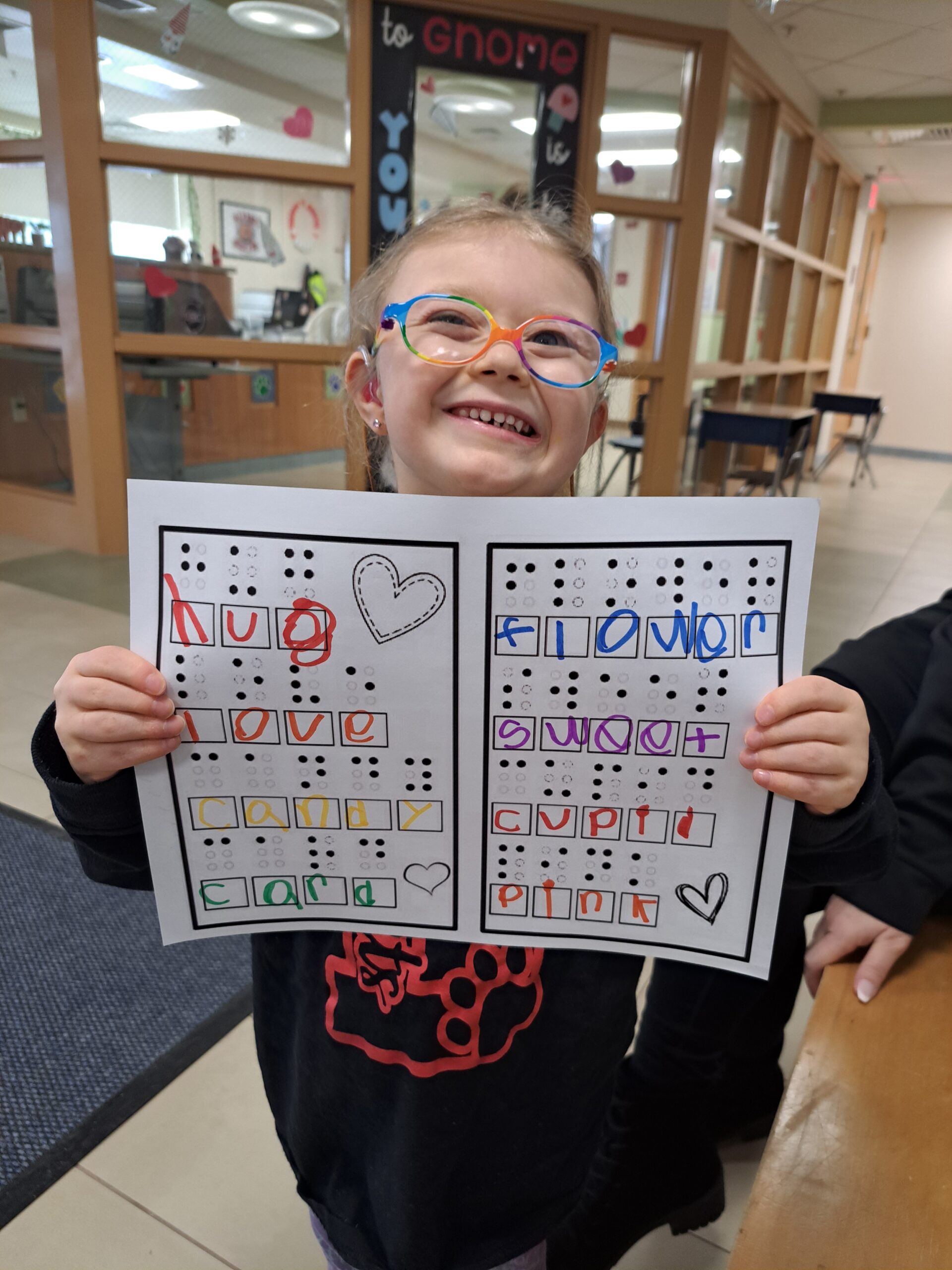A young girl with a huge smile proudly holds up a paper with a braille activity on it.