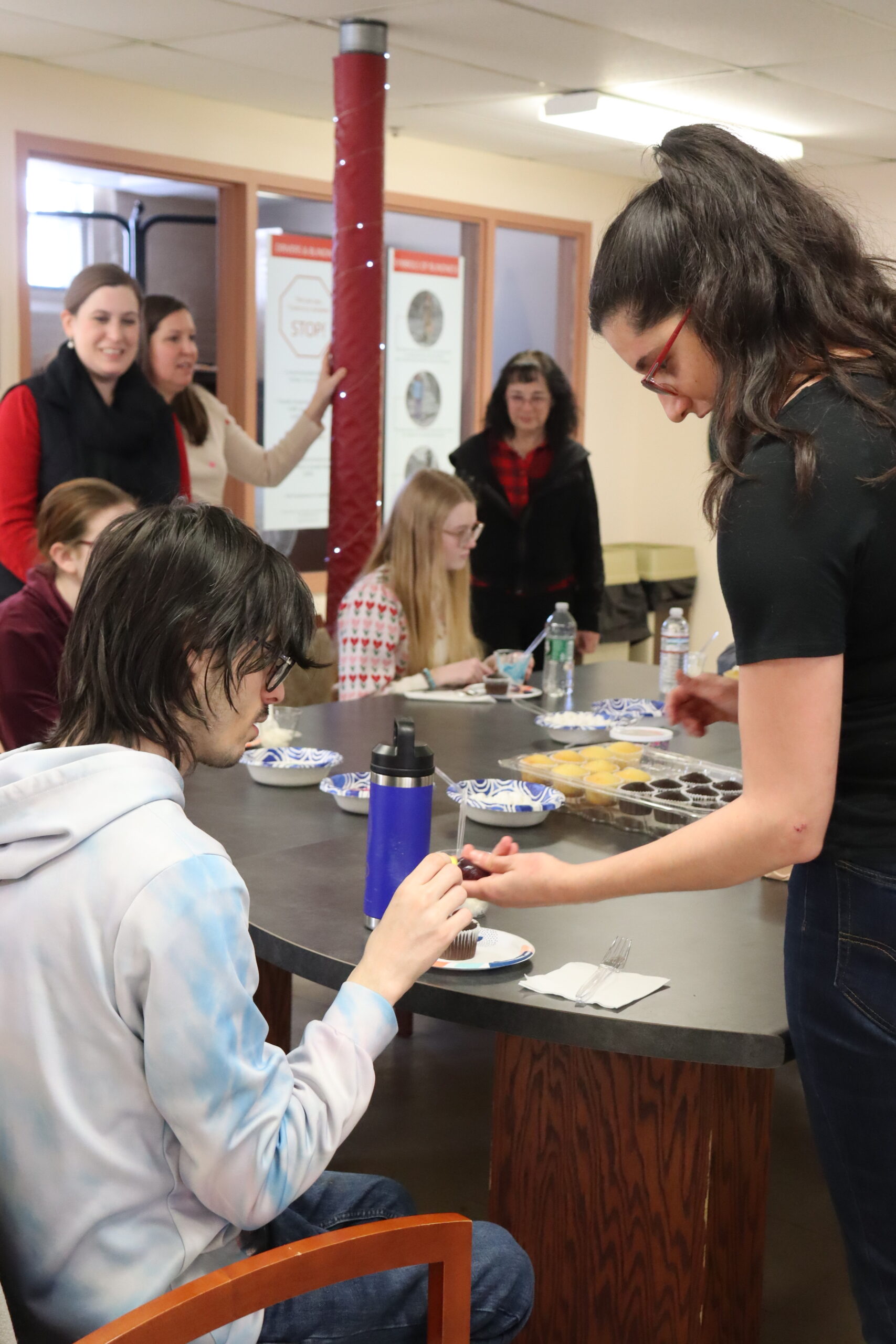 A group of students and teachers sit and stand around a table doing a craft.