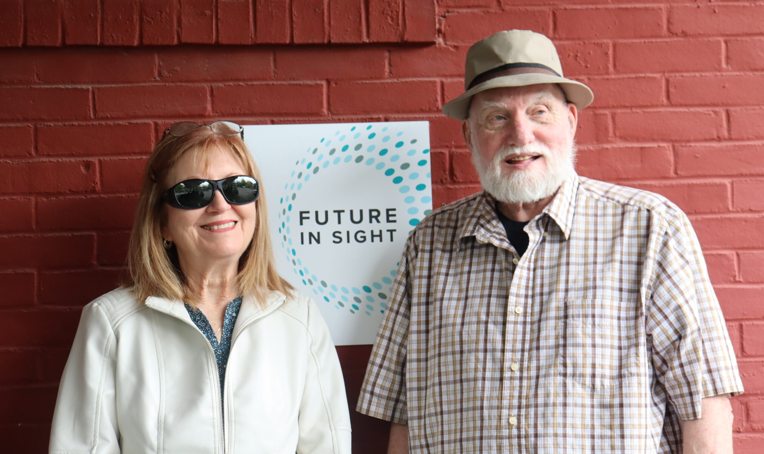 A middle aged woman in sunglasses, a client, poses in front of a Future In Sight sign with a senior man, a volunteer driver.