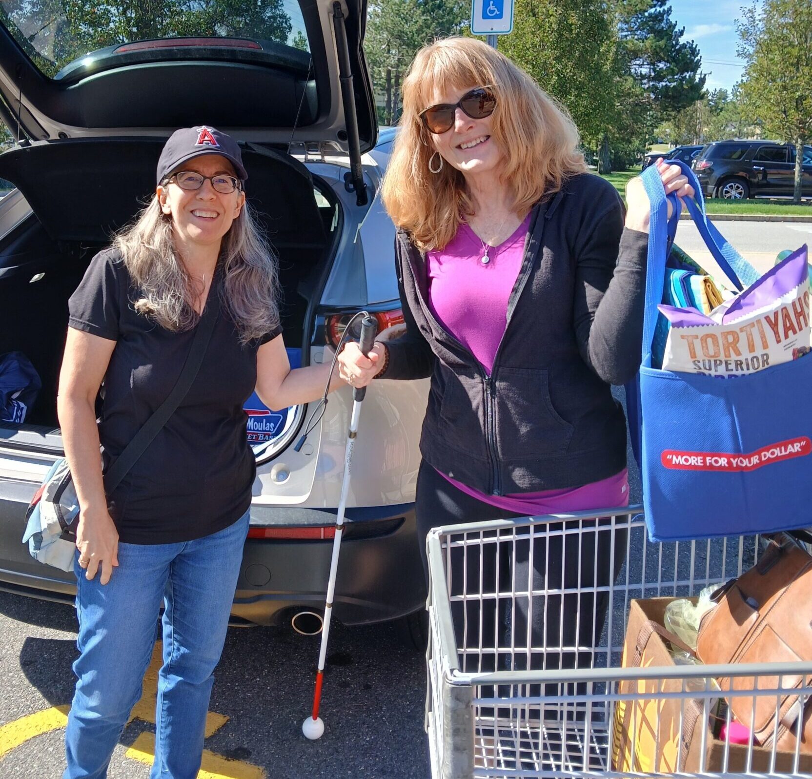 Two women, one with a white cane, load groceries into the trunk of a car.