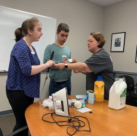 A woman shows a teen girl and boy how to use household items, like an iron.