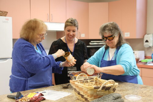 A female instructor and two senior women, one in dark glasses, work together to mix ingredients in a kitchen.