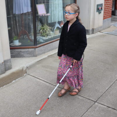 A young girl in glasses walks down the sidewalk using a white cane.