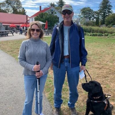 Two individuals, one with a cane and one with a guide dog, outside on a nice day.